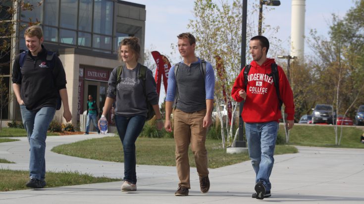Four young students, 3 males and 1 female, walking on the sidewalk in front of the main entrance at the Willmar campus.