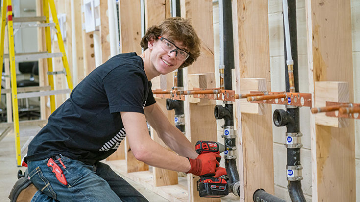 A student plumber smiles at the camera while working on a pipe