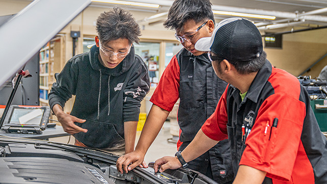 Three auto service tech students working on a car, consulting with each other