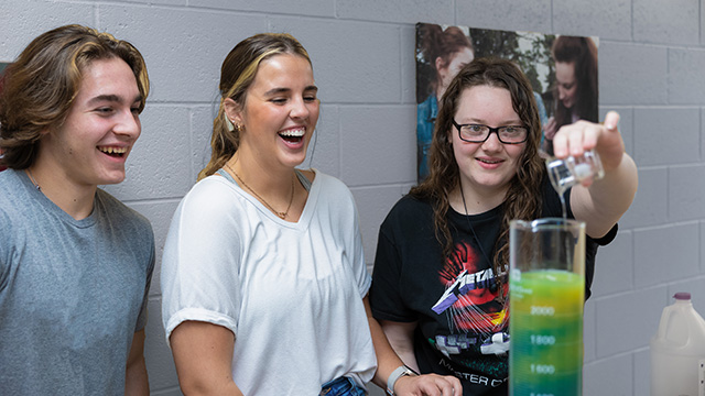 Three students, one on the right is pouring vinegar into a large beaker and the colors of the chemicals are changing and mixing