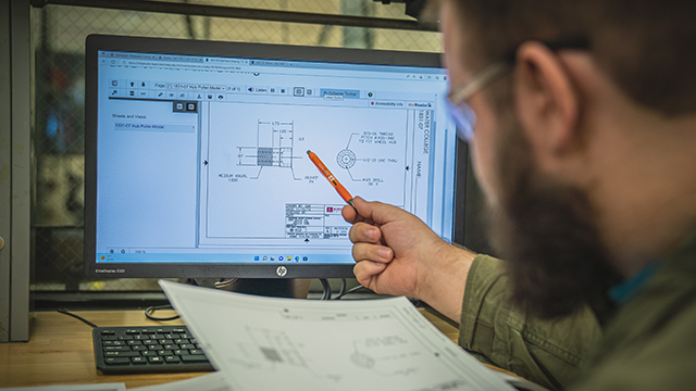 A CADD student, using a pen, gestures towards a computer screen displaying a design while simultaneously holding a paper design in their other hand.
