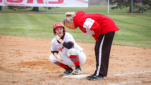 Softball player crouched on the plate, coach bent over listening to the player. Player wearing the RW white uniform and the coach in a red and white Ridgewater jacket
