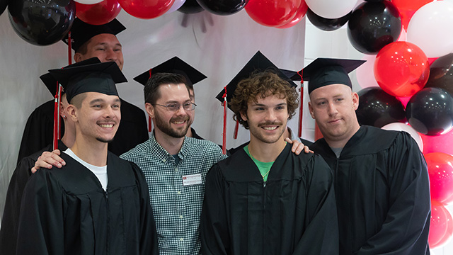 Photo of students in their graduation cap and gowns with NDT instructor. Red, black, and white balloons are behind them.
