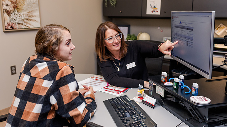 Smiling student on the left, donning a vibrant red Ridgewater t-shirt, holds information, while the recruiter on the right, dressed in a sharp black top and a stylish red blazer, sits with a laptop, both radiating positivity.